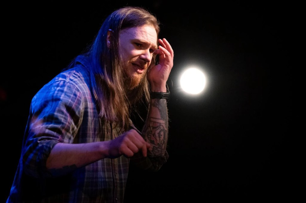 a bearded man with long hair and a plaid shirt. He looks distressed and taps his head with his hand.