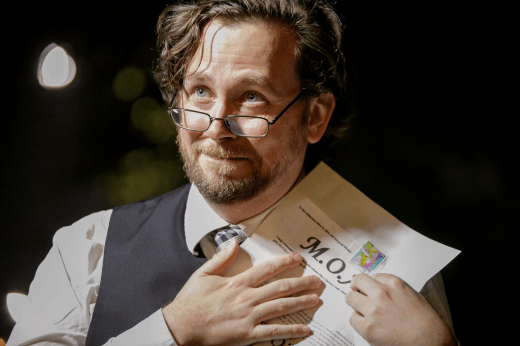 Brian White, a white man in his forties with brown hair and glasses, holds a paper to his chest and smiles warmly.