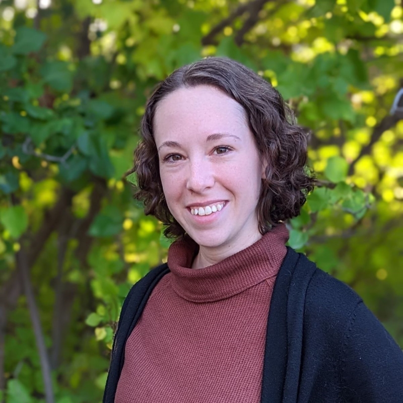 Erin, a woman with short curly brown hair and brown eyes smiling in front of leafy green lilac bushes