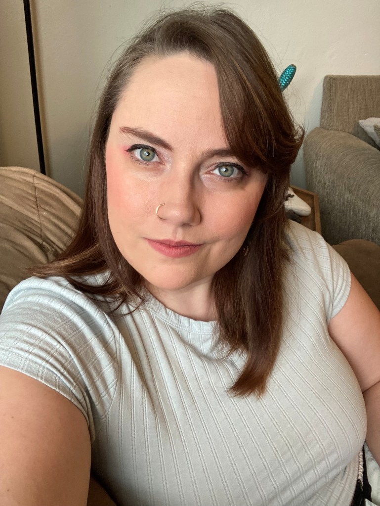 Jasmine, A white woman in her late 20s to early 30s with straight, medium-length brown hair, green eyes, a round face, and a gold nose ring smiles at the camera,