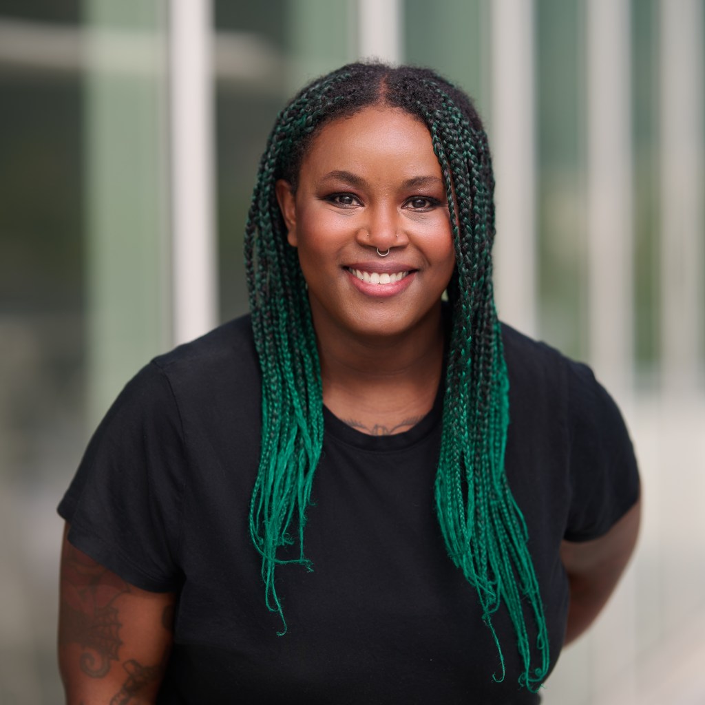 Kateena Bell, a woman with long green box braids leans forward with an easy, confident smile. She has a septum ring, subtle makeup that highlights her eyes, and tattoos on her arms. She wears a simple black t-shirt, and her posture and expression feel open, warm, and grounded