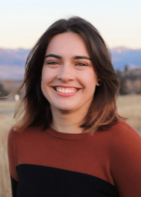Against a starry background, a woman smiles broadly in a headshot. Her shoulder length brown hair blows lightly in a breeze. Text reads Riley Netzley-Hale: Nora
