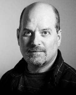 A black and white headshot of a man with a short goatee. He has a slight smile and intense eyes. Text reads James Cox designer
