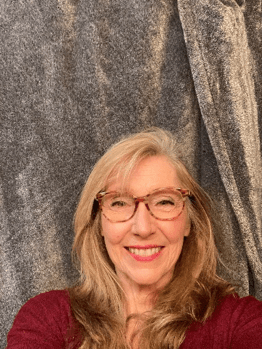 A headshot of an older woman with chest length pale blonde hair. She smiles broadly at the viewer and wears tortoiseshell glasses. Text reads: Ann Peacock: Gatekeeper
