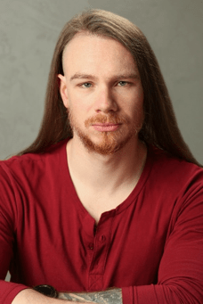 Headshot of Reggie Herbert, a white male in his early thirties. He has long brown hair undercut on the sides, a reddish beard and striking blue eyes. He looks fixedly into the camera with the hint of a smile at the corner of his mouth
